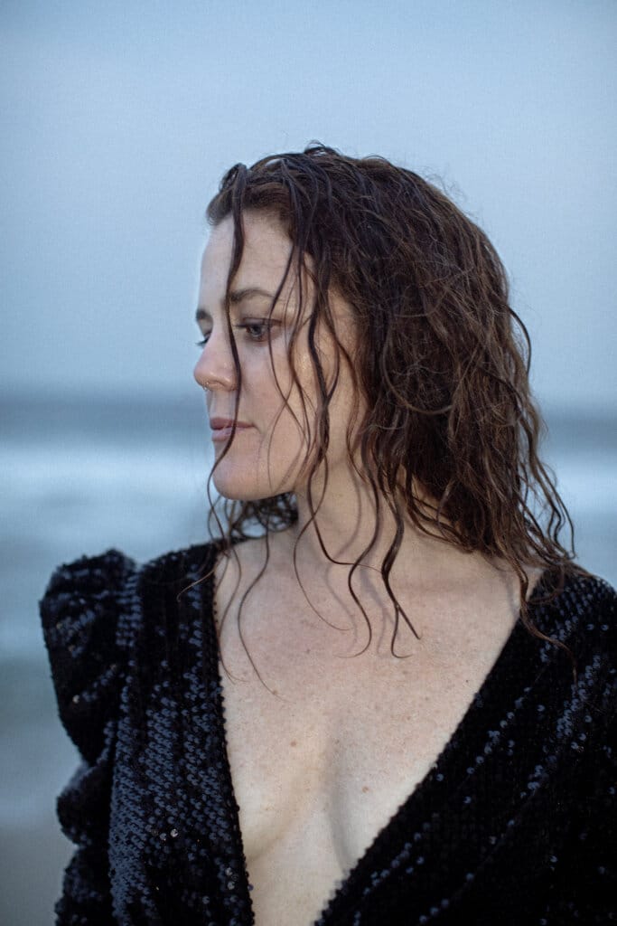Navigating Heartbreak: iskwē's New Single "End Of It All" 2 Promotional photo for "End Of It All" which sees a mid-shot of iskwē wearing a black dress with a v-neckline that reaches her midriff, showing off her cleavage. She has her head turned to the side and her auburn hair is slightly wet. She looks like she's on a cold beach.