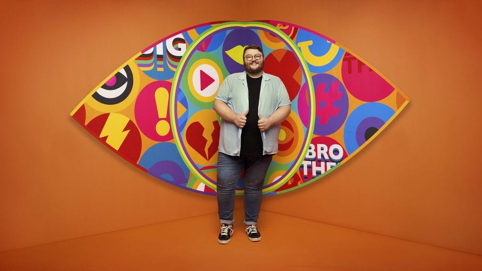 Big Brother UK 2023 Makes a Grand Comeback with Exciting New Housemates and Punishing Games 148 Big Brother 2023 housemate Jenkin standing in front of the new eye logo with a bright orange background.