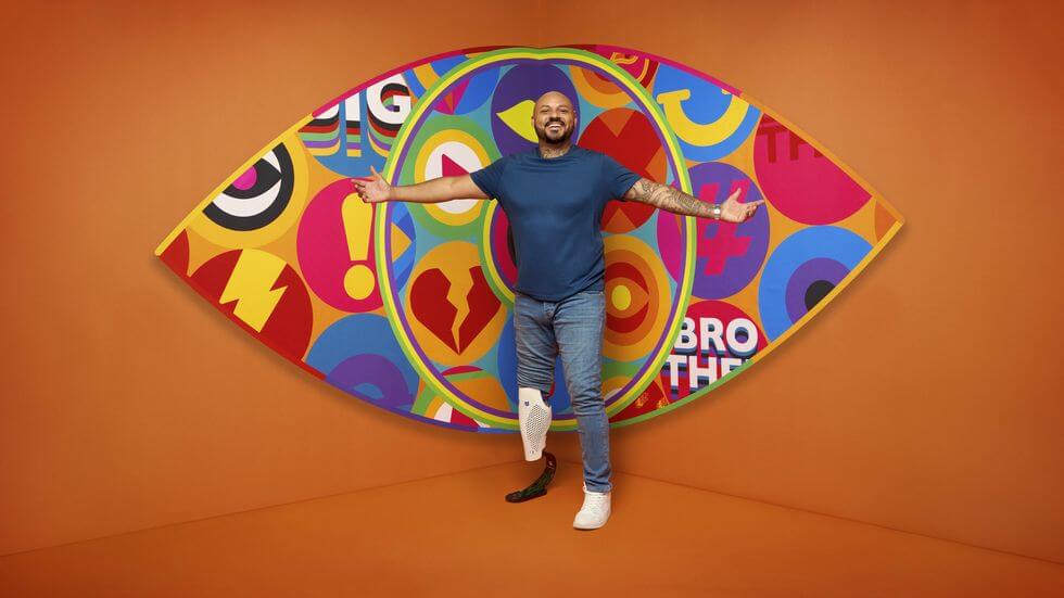 Big Brother UK 2023 Makes a Grand Comeback with Exciting New Housemates and Punishing Games 157 Big Brother 2023 housemate Dylan standing in front of the new eye logo with a bright orange background.