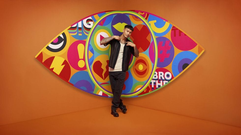 Big Brother UK 2023 Makes a Grand Comeback with Exciting New Housemates and Punishing Games 155 Big Brother 2023 housemate Zak standing in front of the new eye logo with a bright orange background.