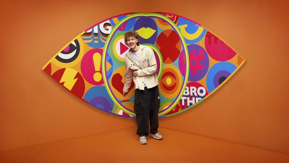 Big Brother UK 2023 Makes a Grand Comeback with Exciting New Housemates and Punishing Games 150 Big Brother 2023 housemate Tom standing in front of the new eye logo with a bright orange background.