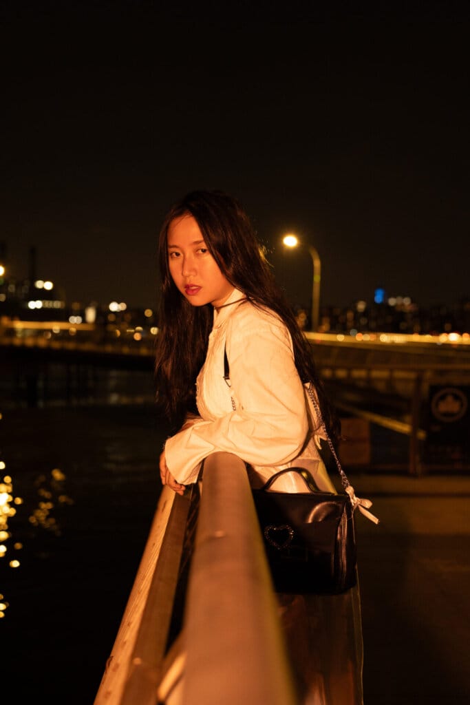 ÊMIA Unveils Sensational Sequel EP "VIDEO CALL: PM" - A Captivating Musical Journey of Emotions and Connection 78 Promotional image for "VIDEO CALL: PM" which sees ÊMIA posing with the New York skyline behind her, leaning against a railing, looking right at the camera.