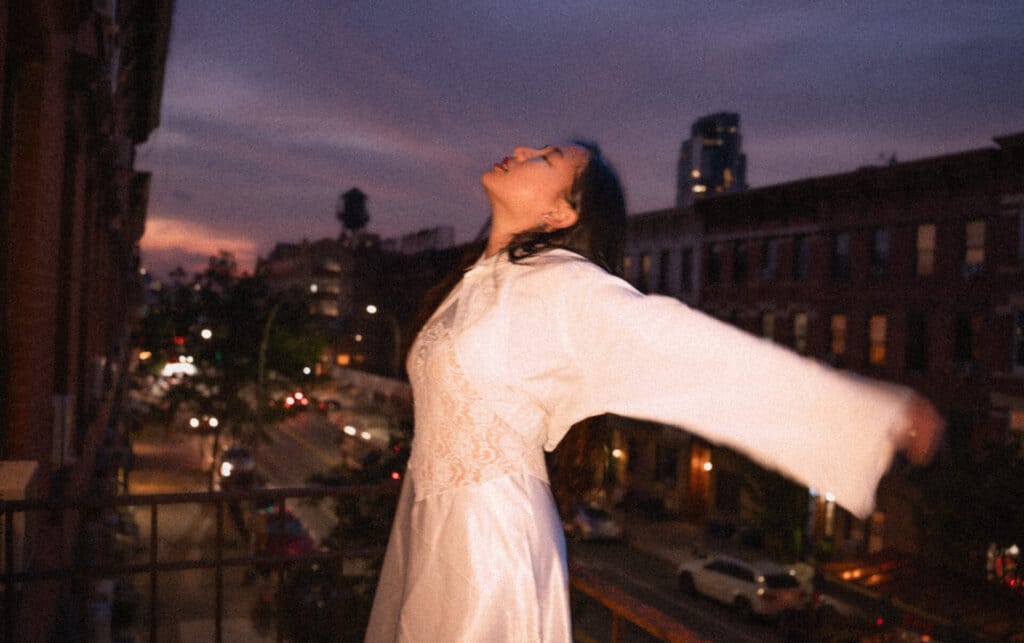 ÊMIA Unveils Sensational Sequel EP "VIDEO CALL: PM" - A Captivating Musical Journey of Emotions and Connection 80 Promotional image for "VIDEO CALL: PM" which sees ÊMIA posing with her arms open wide with the New York skyline behind her as the sun sets or rises.