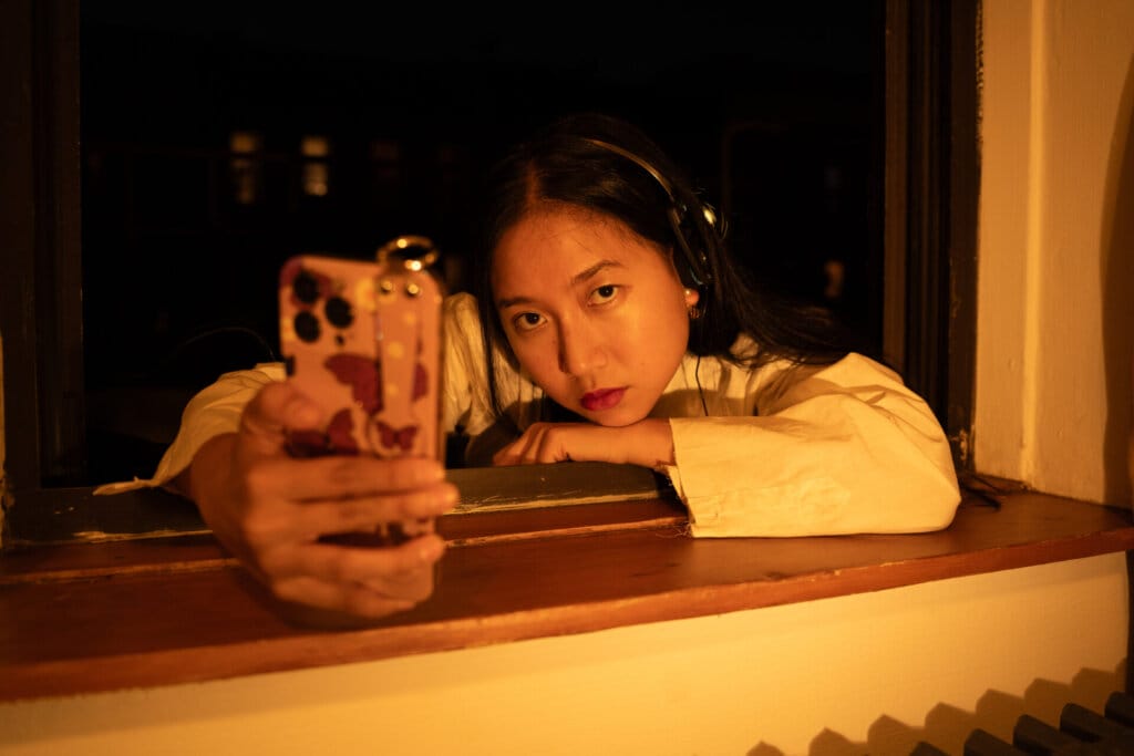 "VIDEO CALL: PM the mini movie": ÊMIA Invites Us into Her World Within A Flawless Visual Masterpiece 35 Promotional image for "VIDEO CALL: PM" which sees ÊMIA posing in a window frame, with her arm perched on the windowsill, looking right at the camera, with her phone out in front of her, taking a selfie.