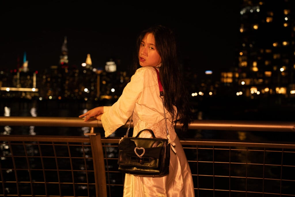 ÊMIA Unveils Sensational Sequel EP "VIDEO CALL: PM" - A Captivating Musical Journey of Emotions and Connection 71 Promotional image for "VIDEO CALL: PM" which sees ÊMIA posing against a rail, with the New York skyline behind her, looking at the camera over her shoulder. She's wearing a white top and a silk skirt with a black bag and it's night time.