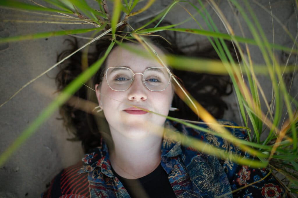 Promotional photo for "Seek The Sun" which sees Clare Siobhan lying down on some sand with a few blades of grass in front of the camera lens.