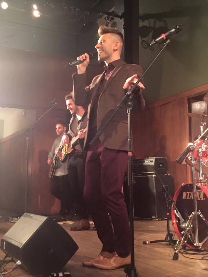 Jay Hofman Unveils Catchy Indie Electro-Pop Earworm "I Can't Sing" 2 Jay Hofman performing on stage, dressed smartly with a burgundy jacket and dark burgundy trousers.