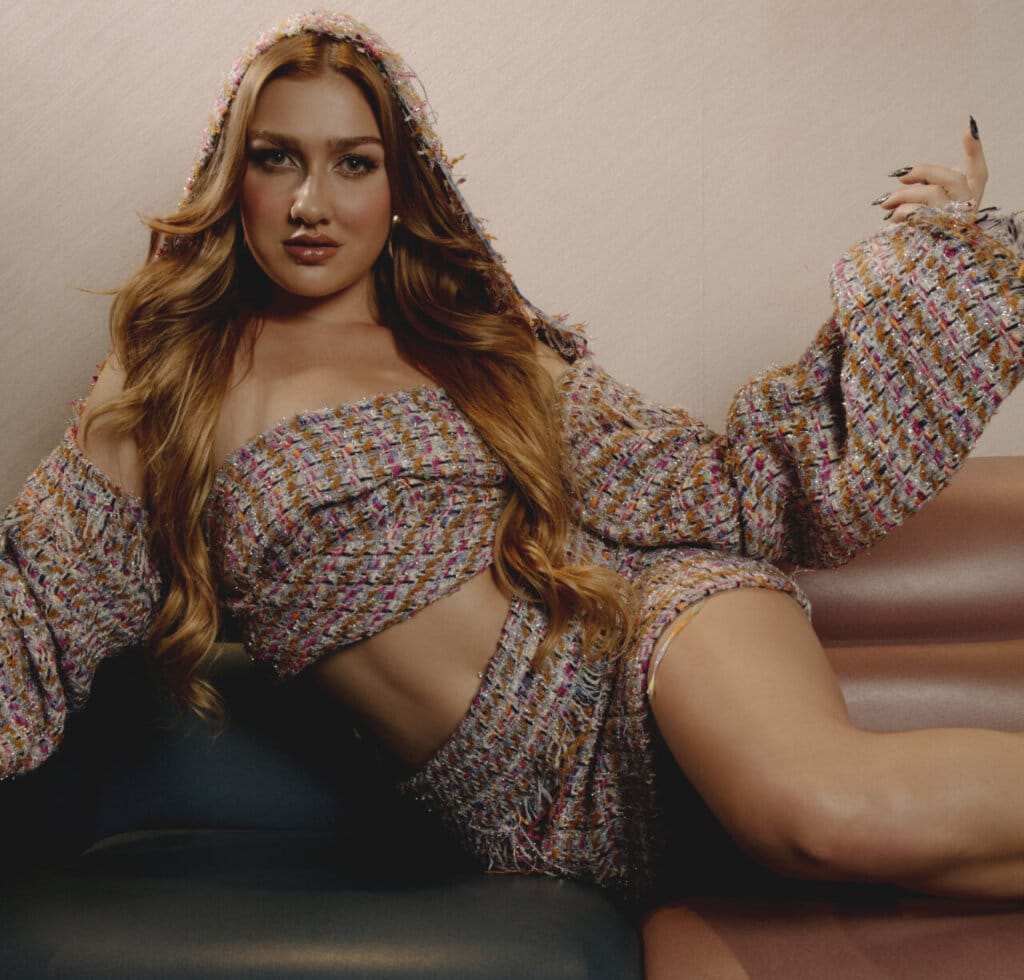 Olivia Lunny lying down on a row of seats that are backed against a wall, wearing a plaid top with matching skirt, resting up on one arm, looking at the camera.