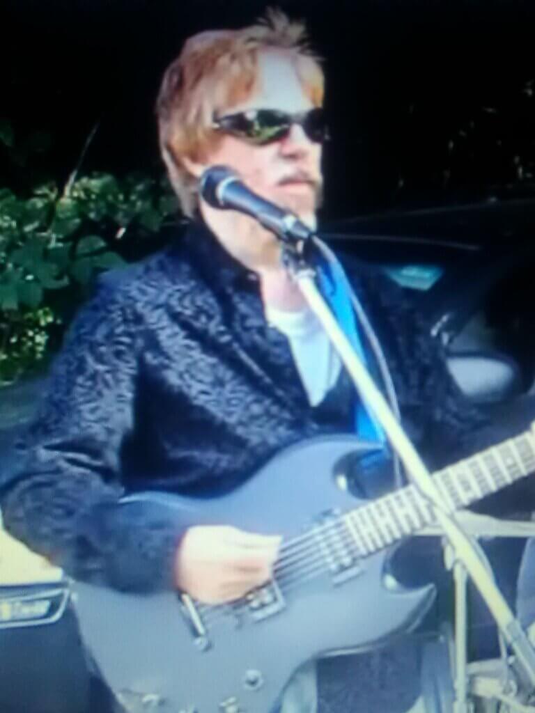 Rich Campanella performing whilst wearing dark shades and holding a guitar in front of a microphone.