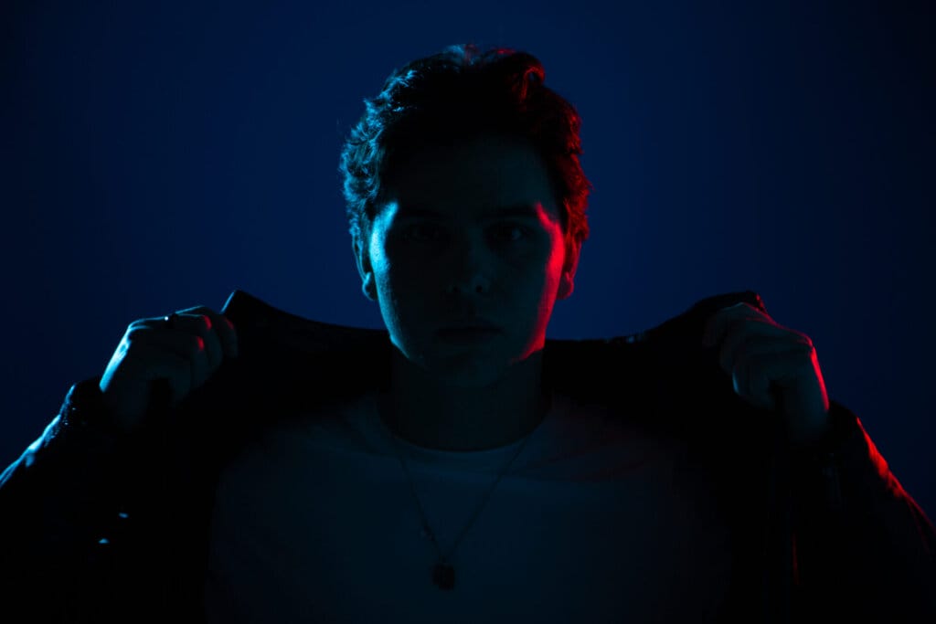 Will Homewood Opens Up: Exclusive Interview on "Burn" and More 58 Will Homewood in the dark, lifting up the collar of his jacket, with a dark blue background. He is mainly covered in shadow.