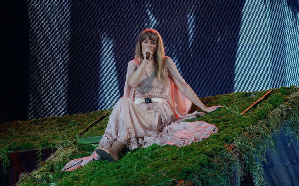 Taylor Swift Postpones The Eras Tour Rio de Janeiro Brazil Show Due to Extreme Heat and Fan Tragedy 36 Taylor Swift sitting on a grassy hill performing on stage wearing a pale pink dress.
