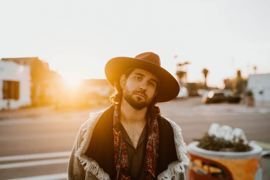 Tyler Del Pino's Provocative Anthem "The Solution" Grapples with Global Struggles 2 Promotional photo for "The Solution" which sees a mid shot of Tyler Del Pino wearing a jacket and a brown fedora hat with a wide brim as the sun sets behind him with rays beaming at the camera and across the road in the background.