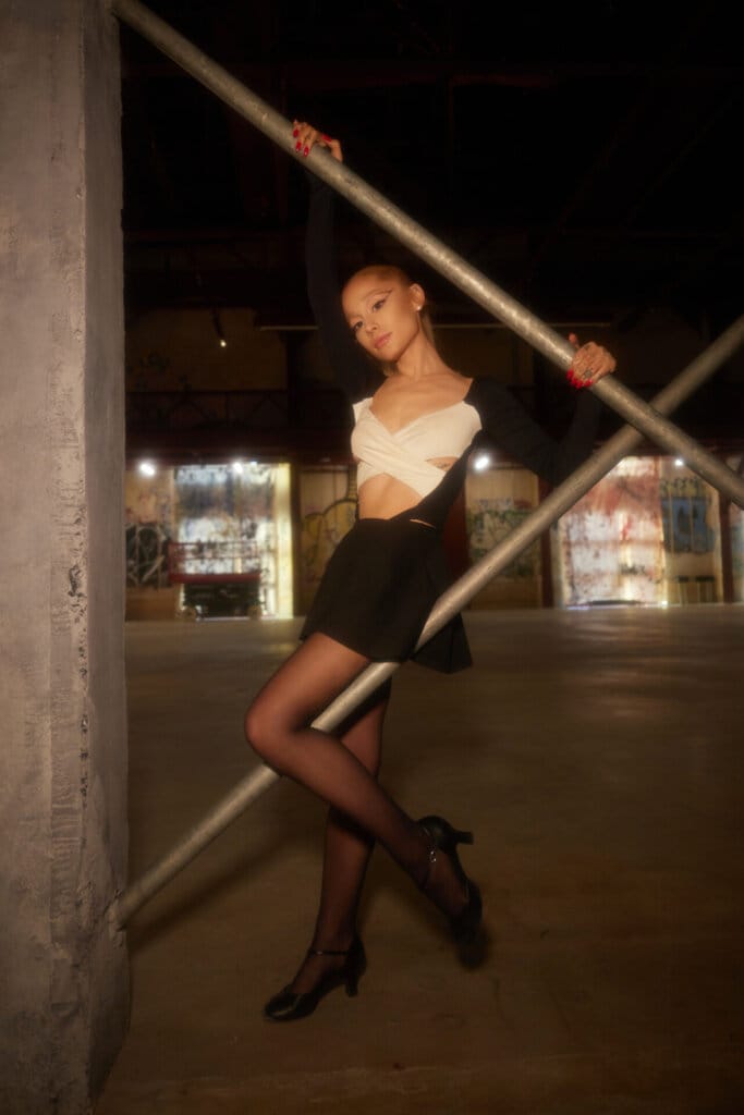 Ariana Grande Unveils Captivating "yes, and?" Single and Music Video 26 Ariana Grande posing for a promotional image for "yes, and?" wearing a black skirt and a white cross top with her blonde hair in a ponytail. She's posing against two grey poles.