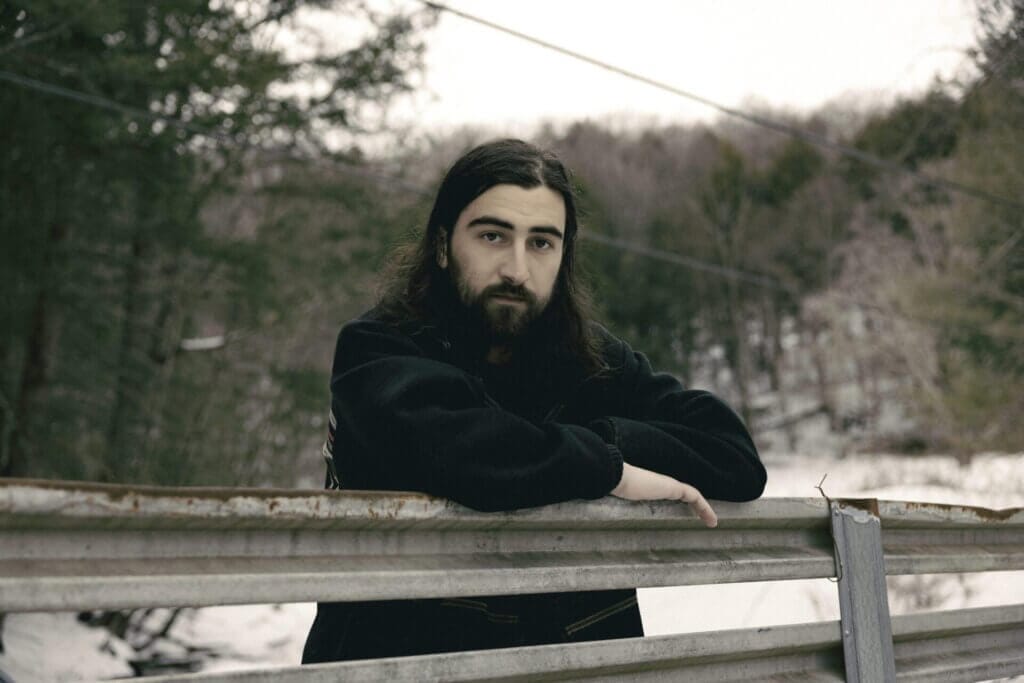 Promotional photo for "Stick Season" which sees Noah Kahan in a black jumper leaning against a wooden fence with his elbows crossed over the top of the fence.