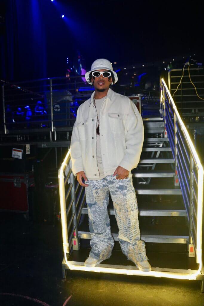 Promotional image of Yung Wylin for "Good Energy" which sees him backstage on the stairs to the stage, posing in a white denim jacket, blue ripped jeans, white trainers, a white hat and white-rimmed shades.