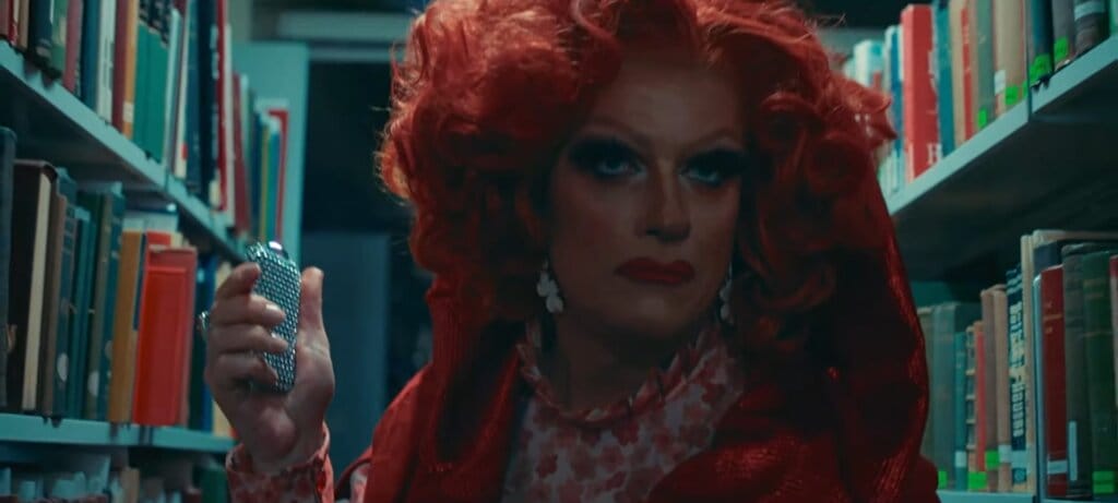 Screenshot from the Wreck series 2 trailer which sees a drag queen with a red wig lurking amongst bookshelves with a bejewelled box in her hand held up as a weapon.