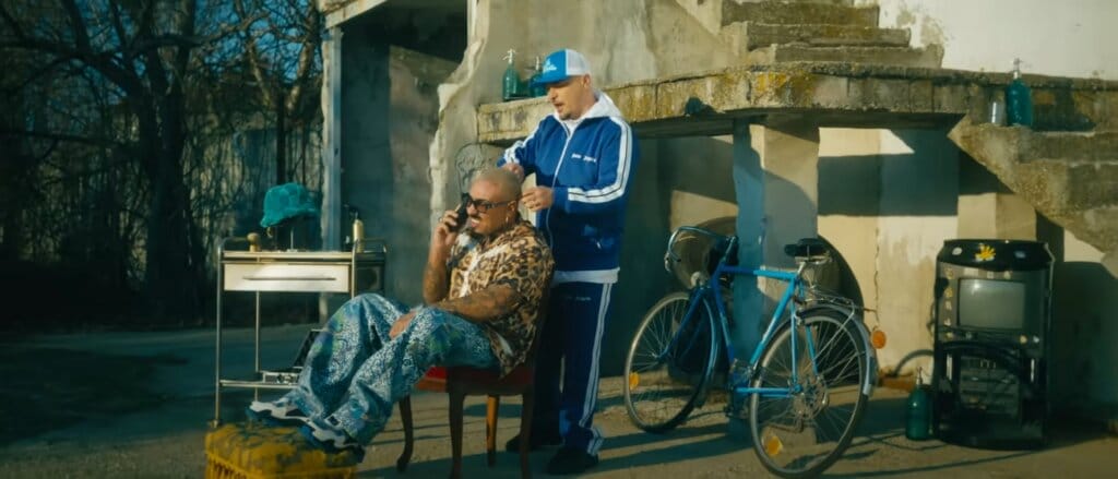 Still from the "Cum se împarte dragostea" music video which sees Puya as a barber dressed in a blue tracksuit as he styles Alex Velea's hair who is sitting on a chair talking on the phone.