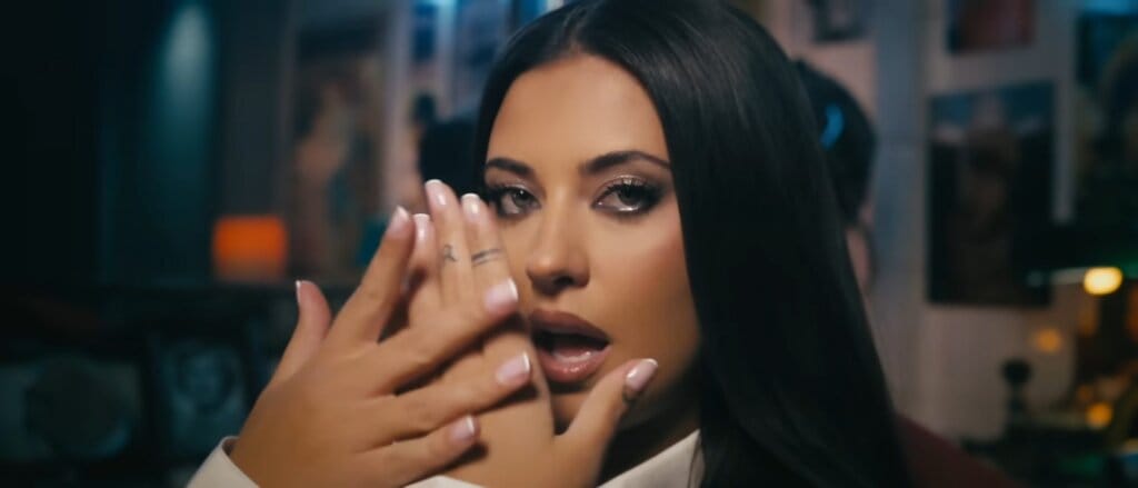 Still from the "Cum se împarte dragostea" music video which sees a close-up image of Antonia slightly hiding her face with her hands as she looks at the camera.