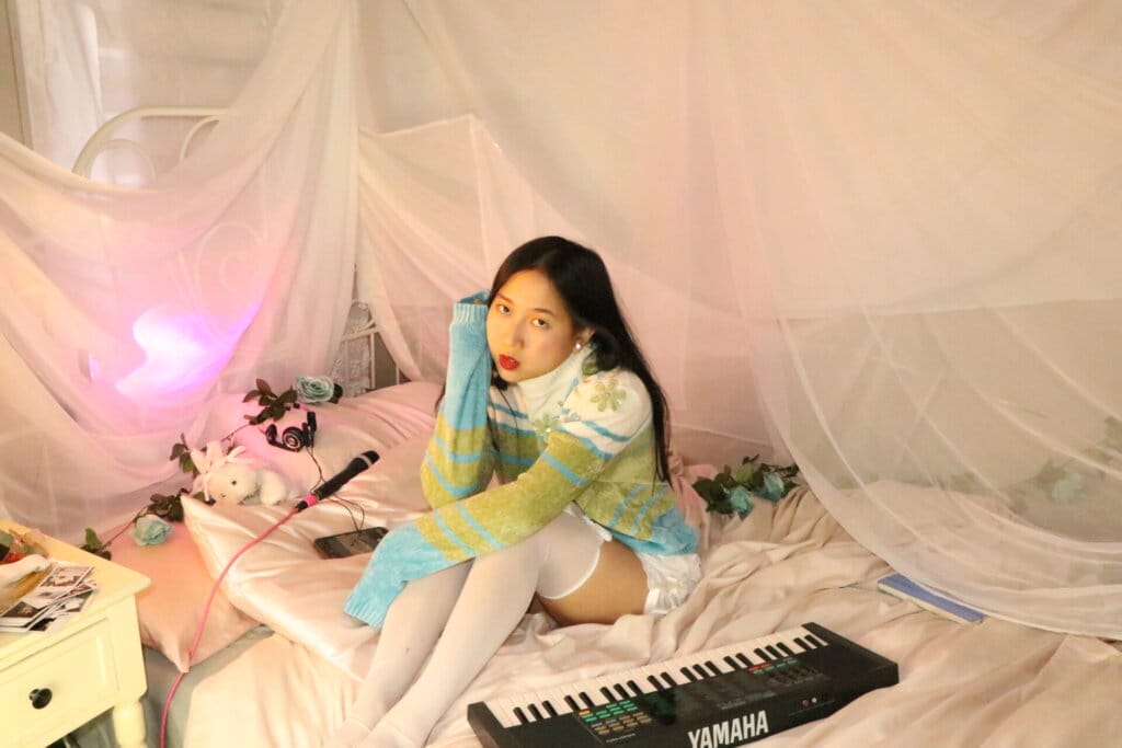 Into the World of ÊMIA: Exploring "Super Fun Party Girl" and Beyond in Our Exclusive Interview With the Indie-Pop Sensation 127 Promotional photo for "Super Fun Party Girl" which sees ÊMIA sitting on a bed with a keyboard out in front of her she hugs her knees, looking at the camera with her dark hair behind her, wearing a white-to-green-to-blue jumper and white stockings. There's a pink hue net over the bed and some small teddies on her pillow.