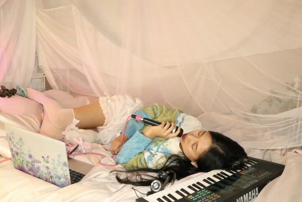 Into the World of ÊMIA: Exploring "Super Fun Party Girl" and Beyond in Our Exclusive Interview With the Indie-Pop Sensation 122 Promotional photo for "Super Fun Party Girl" which sees ÊMIA lying on a bed with a keyboard out in front of her as she rests her head on the keyboard, singing into a microphone with her eyes closed. Her dark hair is on the keyboard and she is wearing a white-to-green-to-blue jumper and white stockings. There's a pink hue net over the bed and some small teddies on her pillow. She has a laptop out in front of her.