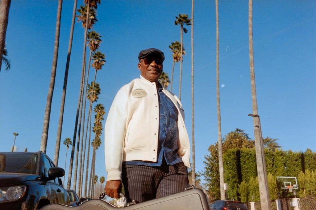 "A Little Bit Of Honey": Marius Billgobenson Soulfully Serenades and Melts Hearts With His New Single 3 Marius Billgobenson standing on what looks like a typical Beverly Hills road, holding a guitar case, wearing a white jacket and a blue shirt as well as some shades.