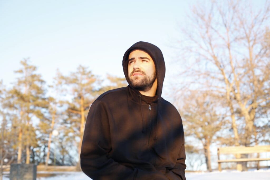 Ben Sefton Explores "Dark Dreams" and Insecurities in His New Single Release 3 Ben Sefton outside wearing a black hoodie with the hood over his head, looking to the left with his hands in the hoodie's pockets, with trees in the distance behind him.
