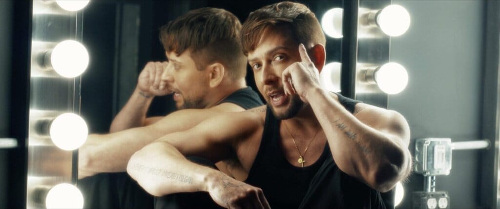 Still from the "I AM" music video which sees David Hernandez wearing a black vest top, leaning against a light-bulb-framed mirror with his knees up and one hand pointing to his forehead in a thinking sign.