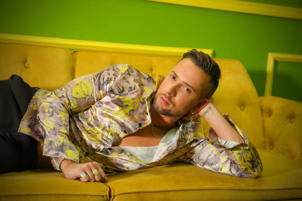 "I AM": David Hernandez Takes Us on an Epic Musical Journey of Growth, Resilience, and Self-Discovery in His New Single and Music Video 20 Promotional photo for "I AM" which sees David Hernandez lying down on a mustard-coloured couch wearing a beige and yellow shirt that is unbuttoned halfway revealing his chest. He's also wearing black jeans. He is stretched out on his side with his elbow on the couch with his hand keeping his head up.