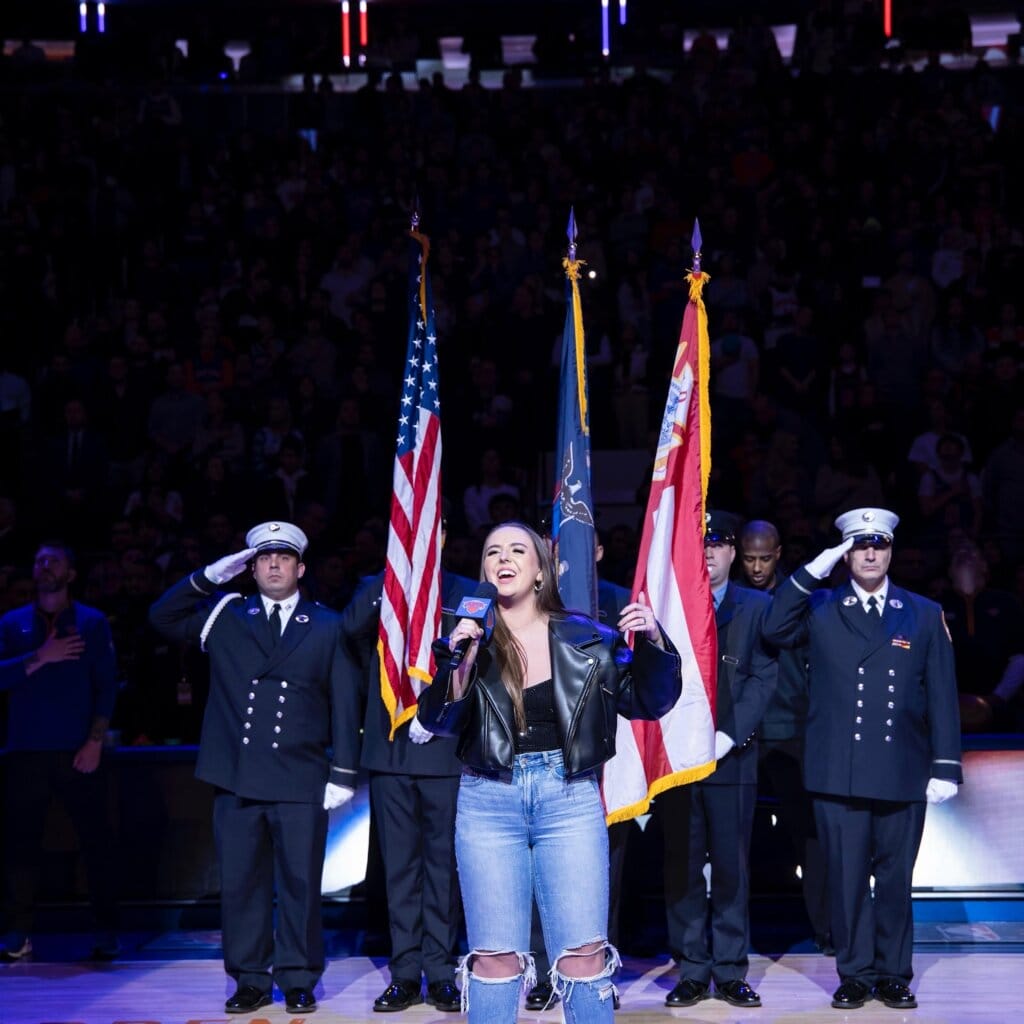 Ali Berke performing the US National Anthem on a stage, wearing knee-cutout jeans, a black top and a black leather jacket. Smartly-dressed men are behind her saluting while there is three flags behind her.