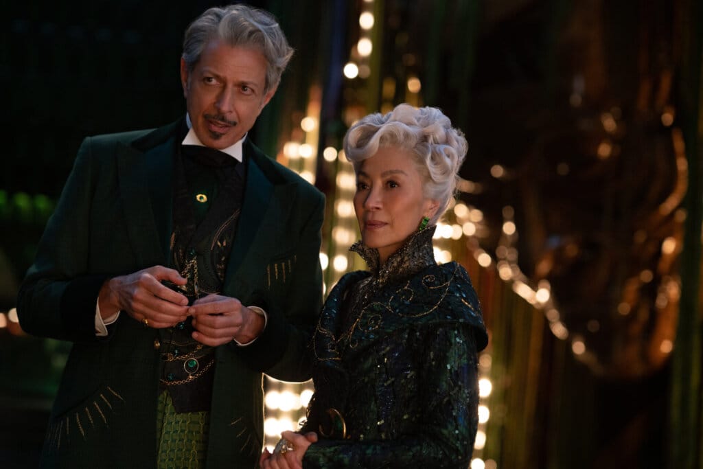 The Wizard of Oz and Madam Morrible clearly discussing someone as they look into the distance past the camera. They're outside at night with multiple lights behind them.