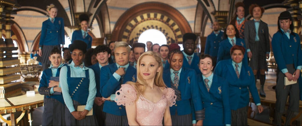 Prepare to Defy Gravity With the Witches of Oz: Wicked Film Trailer Soars with Spectacular Visuals and Unforgettable Performances 20 Glinda in front of a popular crowd who are all wearing blue while she wears pink, while they are in a school setting.