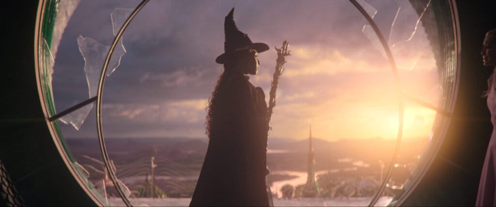 Prepare to Defy Gravity With the Witches of Oz: Wicked Film Trailer Soars with Spectacular Visuals and Unforgettable Performances 23 Elphaba as the Wicked Witch of the West, wearing her black clothes and the pointy hat, holding a broomstick as she flies out a circle window.