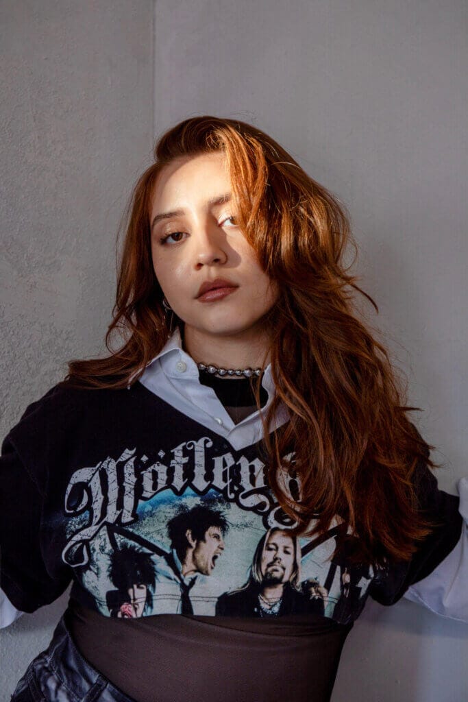 "Time": Maya Ixta Strikes a Perfect Chord With Her Seamless Bilingual Single 12 Promotional image for "Time" which sees a mid-shot of Maya Ixta posing with her hair coming down her right shoulder in curls, wearing a Motley Crew band t-shirt that has been turned into a crop top with a white shirt underneath.