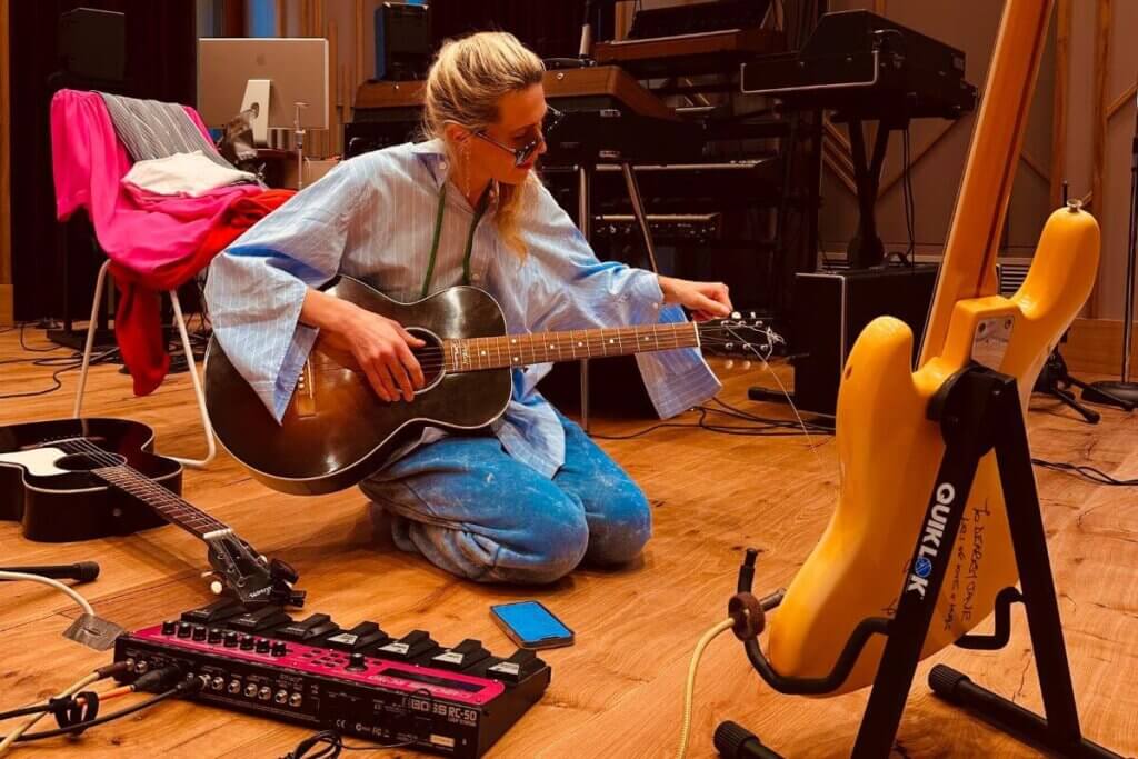 Bird kneeling down in comfy light blue clothes and shades, within a studio with instruments around her, playing the guitar.