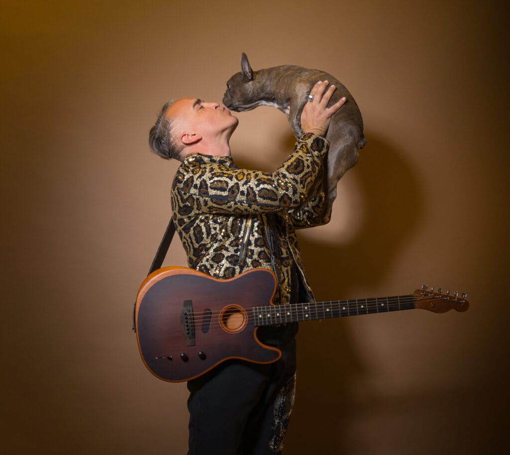 Roger Kuhn and Velvet Code Release New Album "Running With This Dream": Celebrating Identity, Freedom, and Expression 36 Roger Kuhn posing in front of a beige background, wearing a leopard-print shirt and black trousers with a guitar facing us with the strap over his shoulder while he faces to the right, holding a small dog in his hands, held in the air as they kiss.