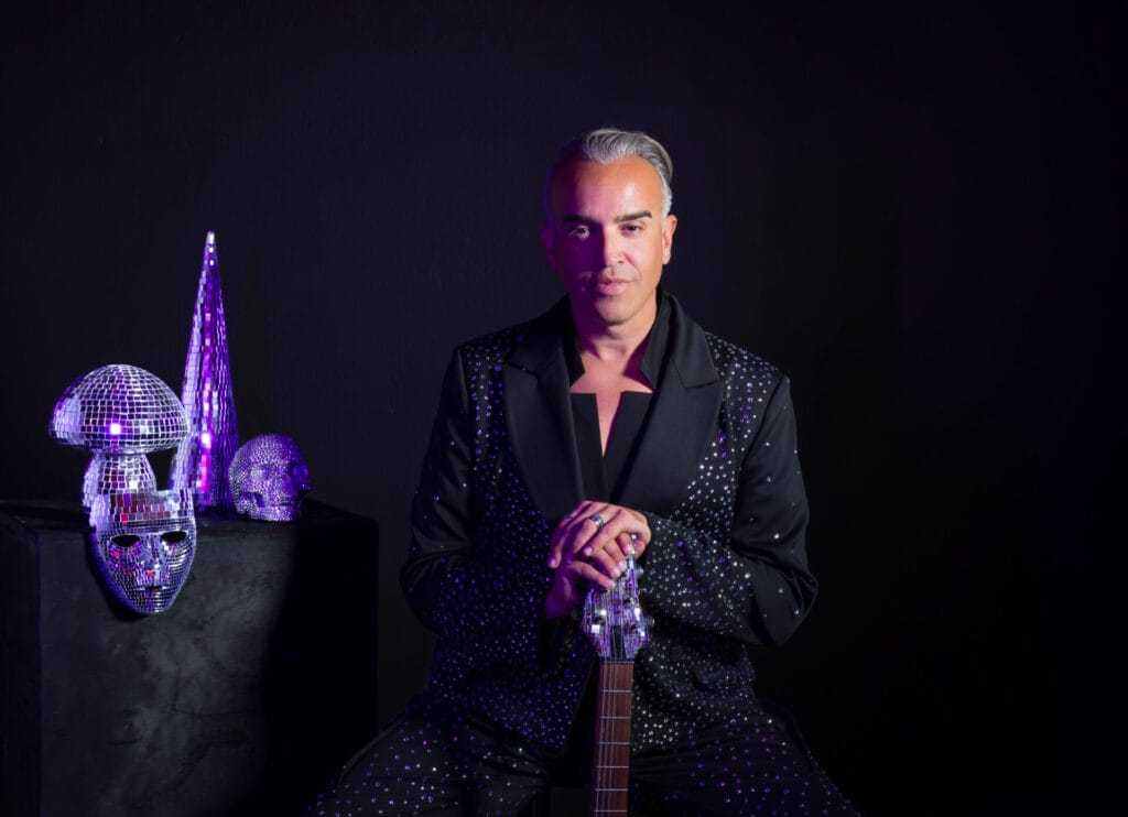 Roger Kuhn posing in a black studio, wearing black with a silver mask to his right and the head of a guitar seen in front of him.