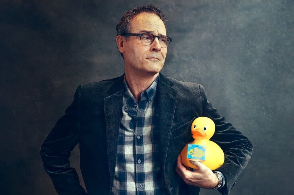 "Send Your River REDUX2": LGBTQ+ Self-Acceptance Celebrated in Keith A. Getchell's Single and Music Video Release 3 Professional photo of Keith A. Getchell standing against a photographer's typical backdrop, holding a rubber duck under his left arm.