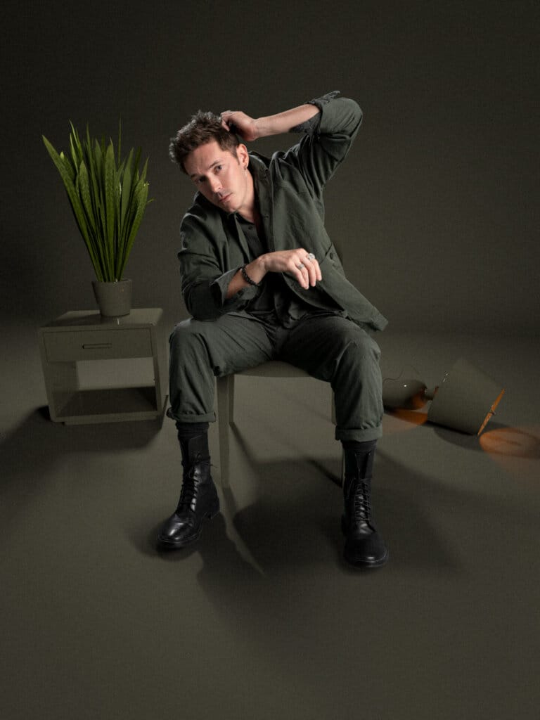 Full length photo of Sam Palladio sitting on s chair, wearing a grey suit with one arm resting on his leg and the other behind his head. There's a plant to the left of him, on a grey table and there's a fallen over lamp to the right.