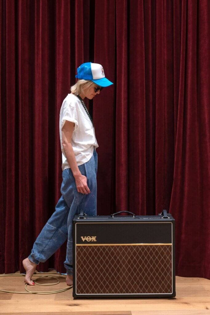 Bird wearing a blue and white snapback cap, a white t-shirt and blue jeans, walking behind an amp, backdropped by a red curtain.