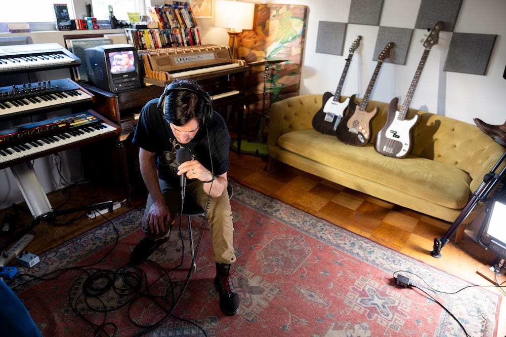 Pete Yorn in the studio, laying down some vocals by singing into a microphone, during his Cardboard Sessions video, where there's a couch behind him with all three cardboard guitars, laying against the wall.