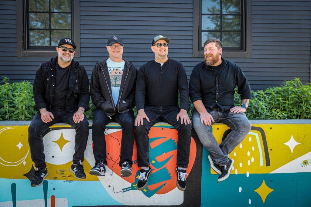 "Ping Pong Ball in a Concrete Room": Names Without Numbers' Anthem for the Overwhelmed 3 Names Without Numbers' four band members sitting on a wall that has art graffiti on it.