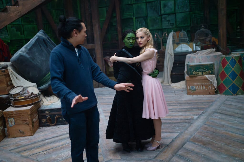 Jon M. Chu directing on the set of Wicked as Ariana Grande and Cynthia Erivo hug. This scene is shown in the featurette.