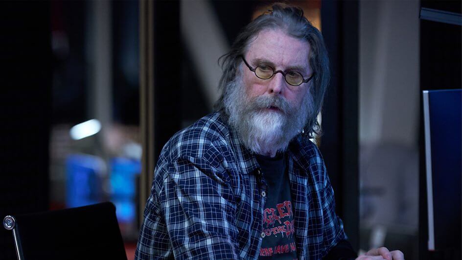 Still from the Nightsleeper trailer which sees an old man with a grey beard (David Threlfall) looking at the camera while wearing a pair of round-shaped glasses.