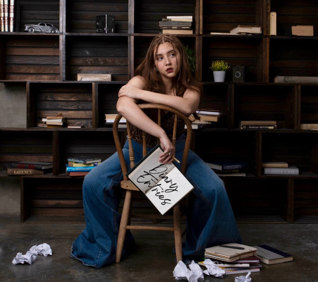 Maya Ixta's Debut EP "Diary Entries": A Heartfelt Journey Through Vulnerability and Resilience 6 Official EP cover artwork for "Diary Entries" which sees Maya Ixta sitting backwards on a chair with a floor-to-ceiling shelfing unit behind her and paper screwed up on the floor. She's wearing a pair of jeans with a huge boot-leg flare at the bottom of them, and she's holding a notebook open that has the title written in calligraphy lettering.