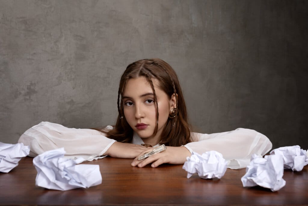 Maya Ixta posing behind a desk which has paper screwed up on, with her arms on the desk and her head just above her arms.