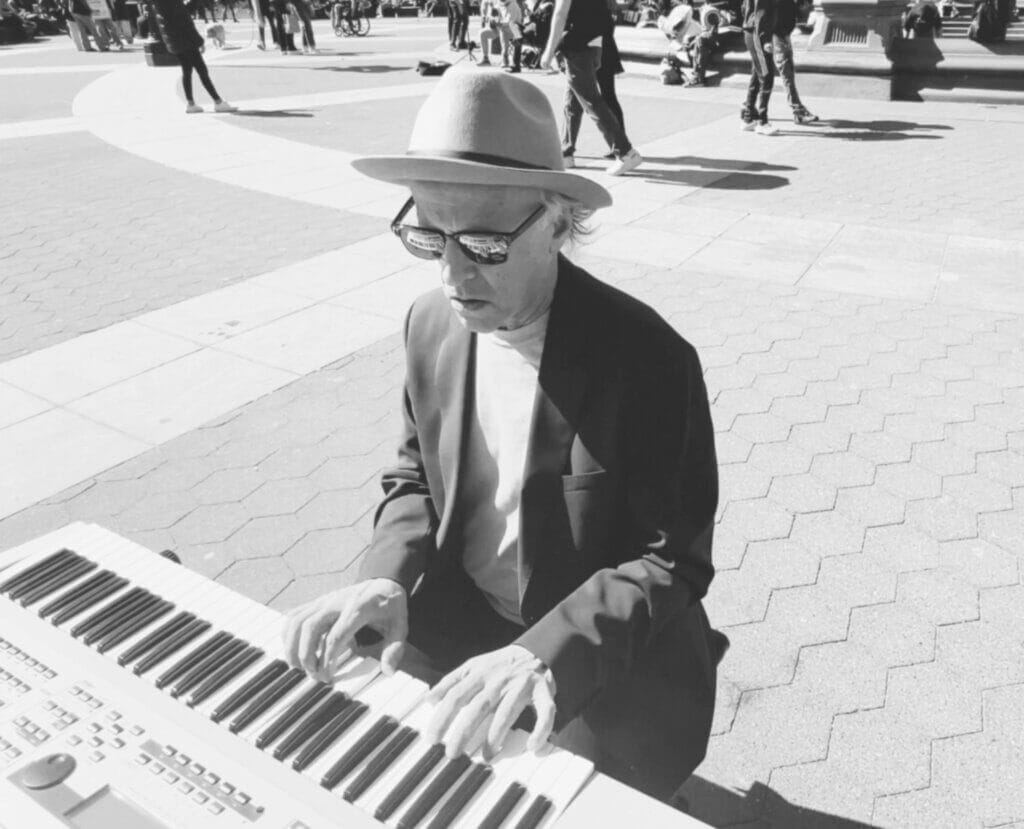 Black and white still from the "When Life's Cold" music video which sees Marshall Oakman playing the keyboard while wearing a hat and sunglasses in Washington Square Park, New York.