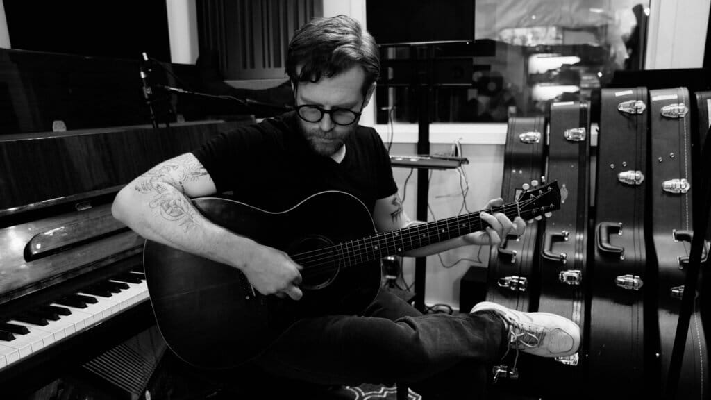 A black and white photo of Michael Burke in the studio sitting at a piano with his back to the keys, playing a guitar.