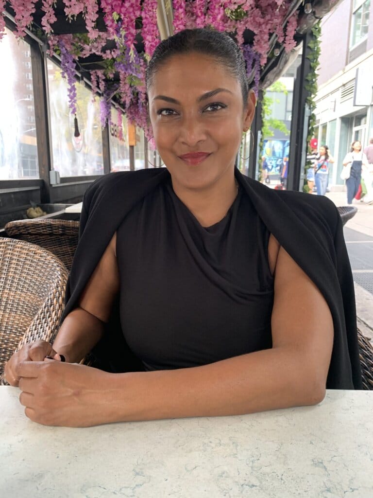 Lauren Atkins, wearing a black dress with a cape that's fitted to her sleeves from her shoulders, sitting at a table with cherry blossoms hanging from the ceiling behind her.