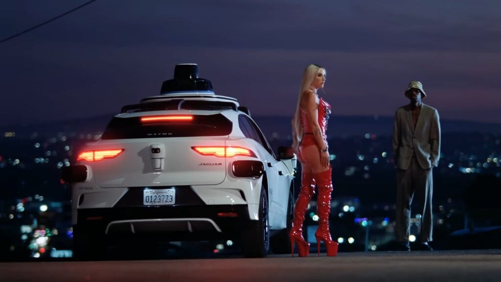 Still from "Joyride" music video which sees Kesha stepping out of a white Jaguar Waymo, dressed in a red leather outfit, and speaking to her bail bondman, in Italian, with a cityscape behind them in the night light.