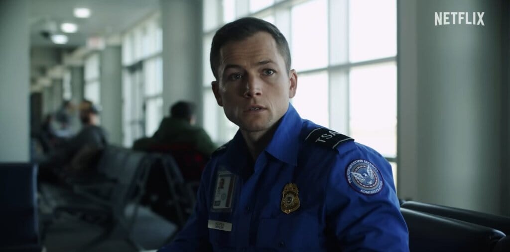 Netflix's Carry-On Epic Film Trailer: Sofia Carson and Taron Egerton Heat Up a Romance That Jason Bateman Turns Into a Suspenseful Thriller 45 Still from Netflix's Carry-On trailer which shows Taron Egerton as Ethan Kopek in TSA agent uniform of a bright blue shirt, sitting down in the airport, chatting with the Mysterious Traveller.